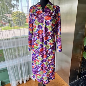 May include: A long-sleeved floral dress featuring a colourful pattern of purple, red, orange, and green. The dress has a collared neckline and a full-length design. The fabric appears to be a lightweight material, suitable for warmer weather.