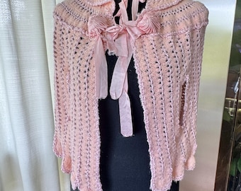May include: A light pink crocheted cape with a scalloped edge and a delicate collar. The cape features a bow and ribbon closure at the neckline, adding a touch of elegance. The open weave design and soft color give it a vintage feel.