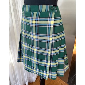 May include: A knee-length wrap skirt featuring a green, blue, and yellow plaid pattern. The skirt has a high waistband with a single dark green button closure on the right side and vertical pleats along the side and back sections.