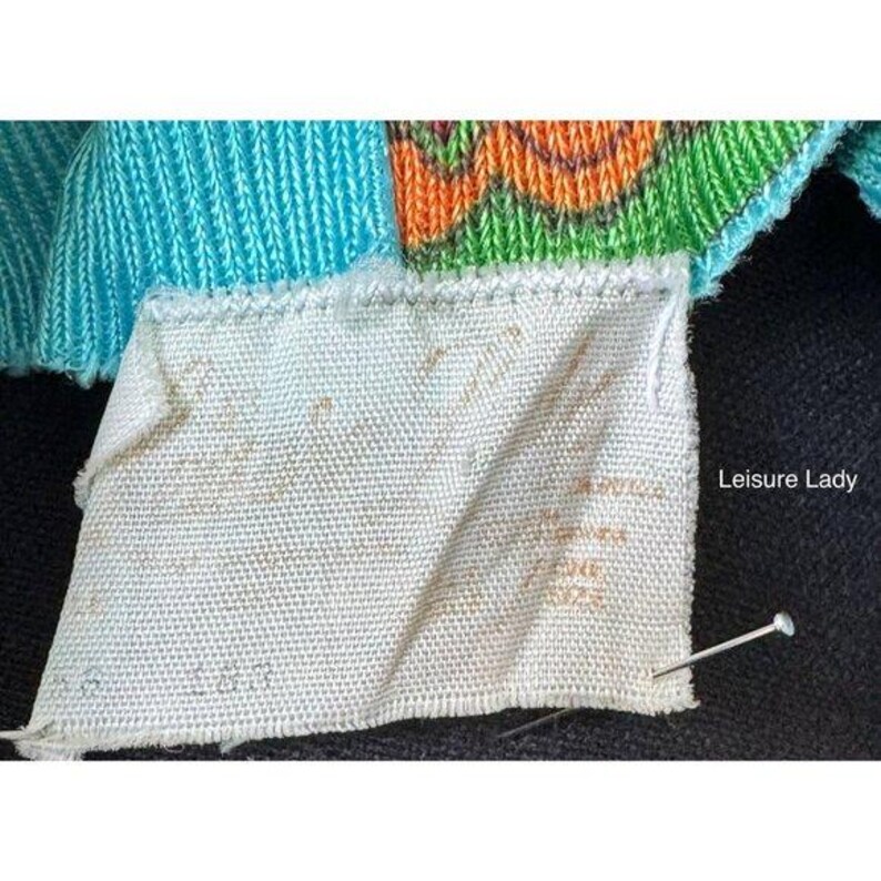 Puede incluir: Una etiqueta de ropa de tela blanca con texto desvanecido unida a una prenda de punto con secciones en turquesa, naranja y verde. Un alfiler recto plateado est&aacute; insertado en la esquina inferior derecha de la etiqueta sobre una superficie oscura.