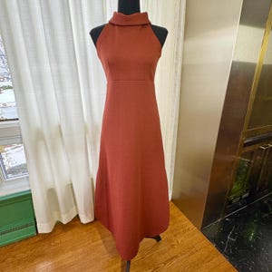May include: A rust-colored, sleeveless maxi dress with a high neckline. The dress is displayed on a mannequin, showcasing its A-line silhouette. The fabric appears to have a subtle texture. The dress is floor-length.