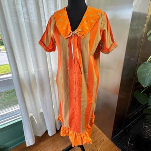 以下が含まれることがあります： オレンジとタンのストライプ柄のドレス。大きなオレンジ色の襟に黄色の水玉模様。ドレスは半袖で、裾はフリル。首元には白いリボン。黒いマネキンに着せられています。