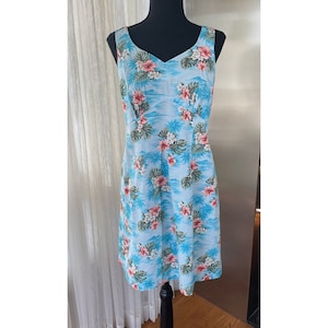 May include: A sleeveless, light blue dress with a V-neckline. The dress features a tropical floral print with pink and white flowers, green leaves, and palm trees. The dress is displayed on a black mannequin.