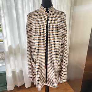 Könnte beinhalten: Ein Cape mit Hahnentrittmuster in Beige, Schwarz und Weiß, auf einer Schaufensterpuppe präsentiert. Das Cape hat einen hohen Kragen, eine Frontöffnung und zwei Vordertaschen. Der Stoff hat ein klassisches Webmuster.