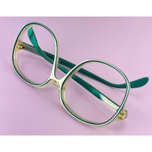 May include: Oversized eyeglasses featuring a two-tone design with a translucent pale yellow front frame and deep emerald green inner rims and temples. The frames have a rounded, wide shape with a high bridge and thin, lightweight construction.