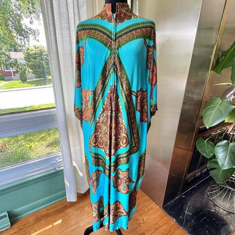 Puede incluir: Un caft&aacute;n largo hasta el suelo con un color base turquesa vibrante y un intrincado patr&oacute;n sim&eacute;trico de cachemira y flores en tonos naranja, dorado y verde oscuro. La prenda presenta un cuello alto con un cierre de cremallera central.