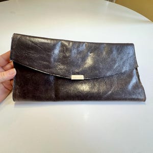 May include: A dark brown leather clutch with a flap closure and a silver-colored rectangular clasp. The clutch is rectangular and appears to be made of distressed leather. The flap folds over the top of the clutch.