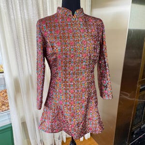 May include: A long-sleeved dress featuring a high mandarin collar and a vibrant, multi-colored metallic plaid pattern. The fabric displays a textured, shimmering weave with interwoven threads of pink, orange, green, and gold, creating a structured silhouette.