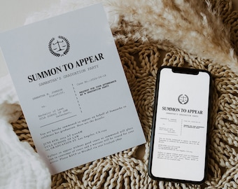 May include: A white card with the text "SUMMON TO APPEAR" and "SAMANTHA'S GRADUATION PARTY" in black text. The card is on a woven mat with a bouquet of dried pampas grass. A smartphone with a similar card displayed on the screen is also on the mat.