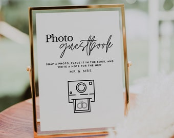 May include: A gold-framed sign with the text "Photo guestbook" in black cursive font. The sign instructs guests to snap a photo, place it in the book, and write a note for the new Mr. & Mrs. A black and white illustration of a camera with two rings inside is below the text.