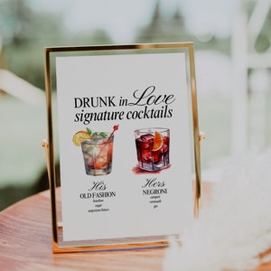 May include: A gold-framed sign with the text "DRUNK in Love signature cocktails" and illustrations of two cocktails. The text below the illustrations describes the ingredients for each cocktail: "His OLD FASHION bourbon, sugar, angostura bitters" and "Hers NEGRONI campari, vermouth, gin".