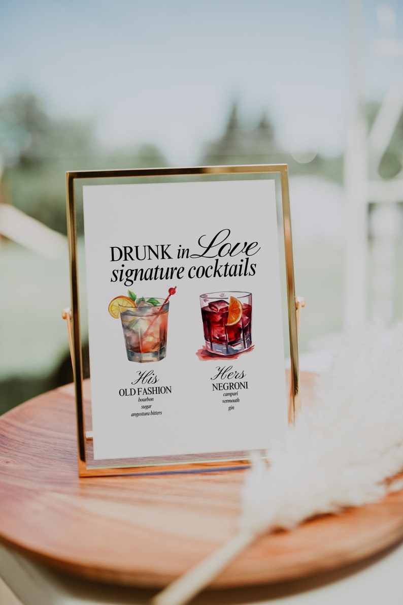 May include: A gold-framed sign with the text "DRUNK in Love signature cocktails" and illustrations of two cocktails. The text below the illustrations describes the ingredients for each cocktail: "His OLD FASHION bourbon, sugar, angostura bitters" and "Hers NEGRONI campari, vermouth, gin".