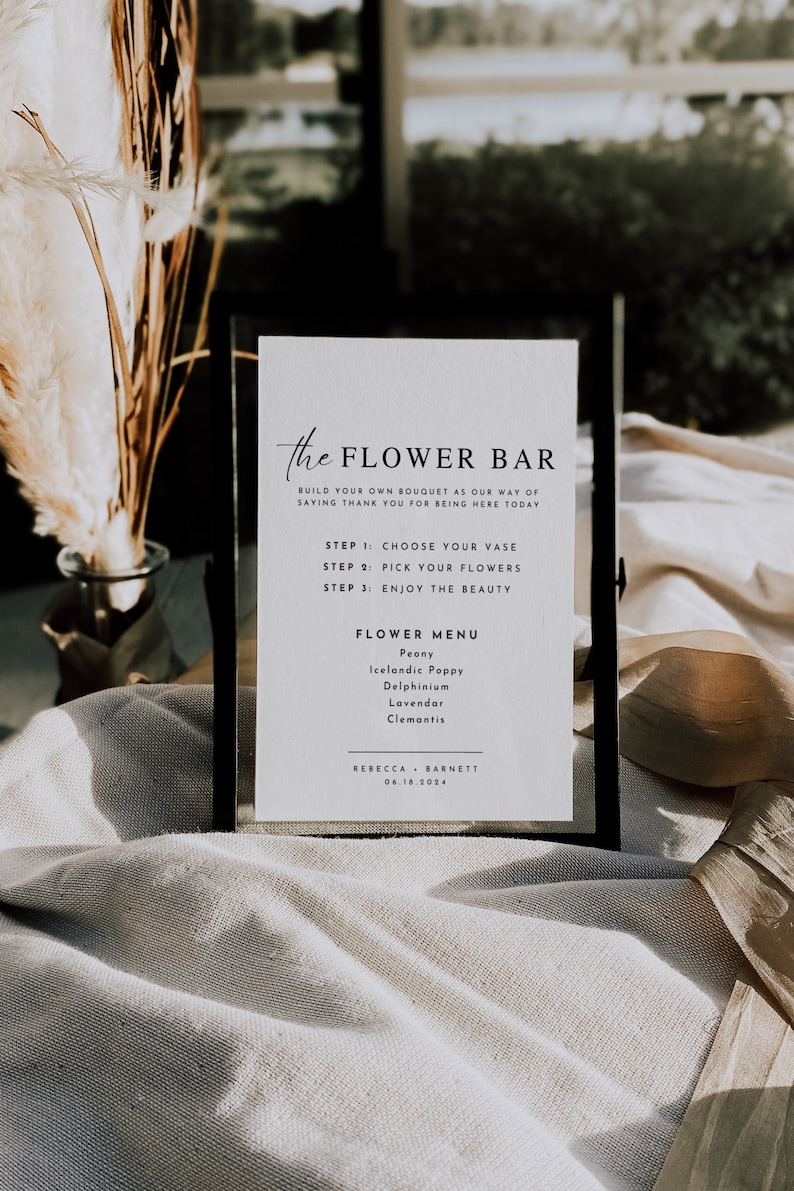 May include: A white sign with black text that reads "the FLOWER BAR" and instructions for building your own bouquet. The sign also includes a flower menu with the following options: Peony, Icelandic Poppy, Delphinium, Lavendar, and Clemantis. The sign is in a black frame and is sitting on a white fabric surface.