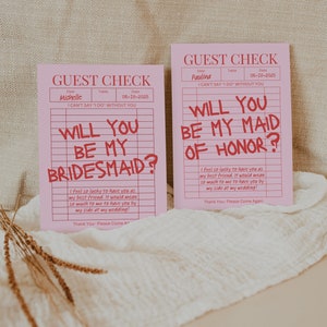 May include: Two pink guest check cards with the text "Will you be my bridesmaid?" and "Will you be my maid of honor?" written in red. The cards have a grid pattern and the text "I can't say 'I do' without you" is printed on both cards.
