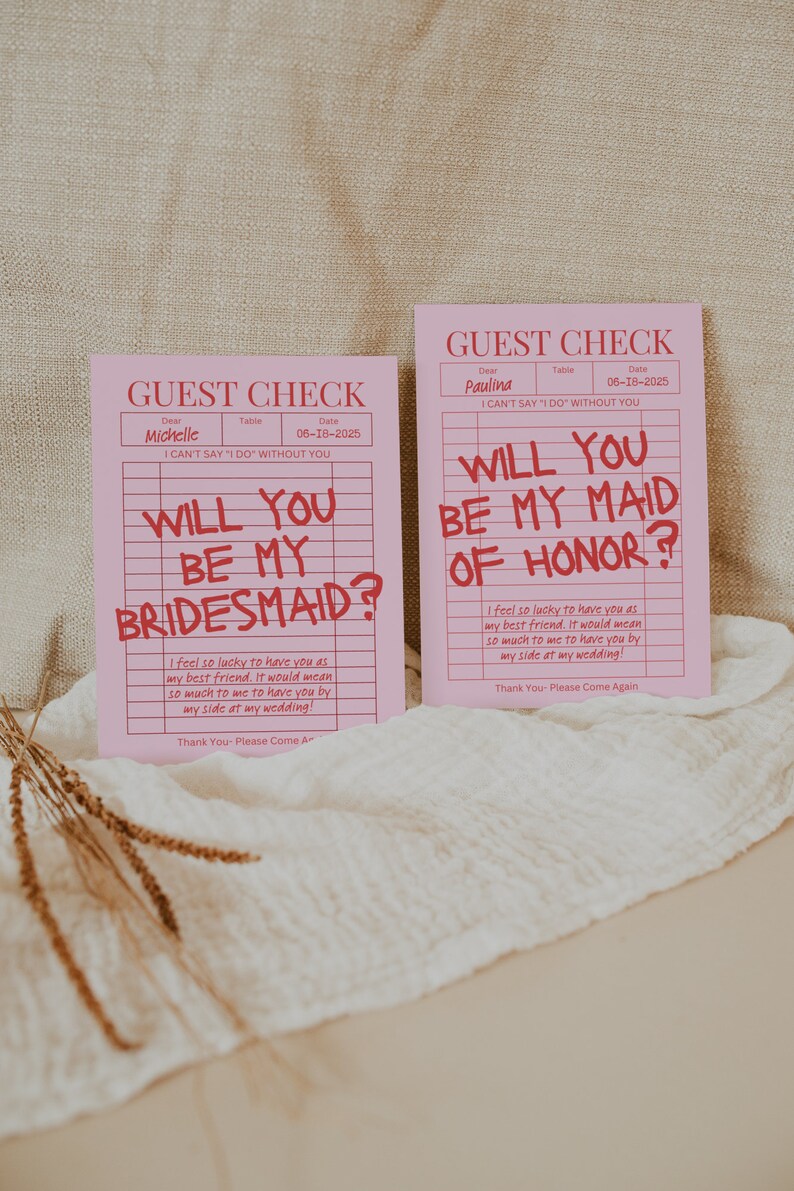 May include: Two pink guest check cards with the text "Will you be my bridesmaid?" and "Will you be my maid of honor?" written in red. The cards have a grid pattern and the text "I can't say 'I do' without you" is printed on both cards.