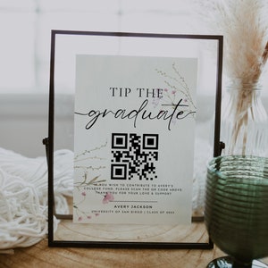 May include: A white sign with a floral design and the text "Tip the graduate". A black and white QR code is below the text. The sign is in a black frame and is sitting on a wooden surface.