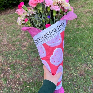 May include: A bouquet of pink and white flowers wrapped in pink and white paper with a heart pattern. The paper has the text "The Valentine Times" and "Wednesday, February 14, 2024" printed on it.