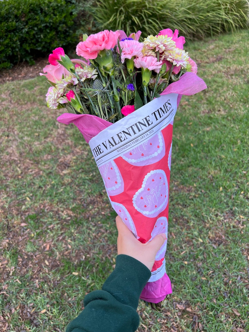May include: A bouquet of pink and white flowers wrapped in pink and white paper with a heart pattern. The paper has the text "The Valentine Times" and "Wednesday, February 14, 2024" printed on it.
