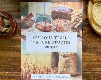 May include: A book titled "Curious Trails Nature Studies Wheat" with images of bread, wheat, and flour. The book is on a wooden surface with a rolling pin and a loaf of sliced bread. The book's cover features the text "By Books and Willows."