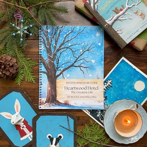 May include: A winter-themed still life featuring a book titled "Heartwood Hotel The Greatest Gift" with a snowy tree illustration. The scene includes a teacup with a lit candle, decorative art, and holiday greenery.