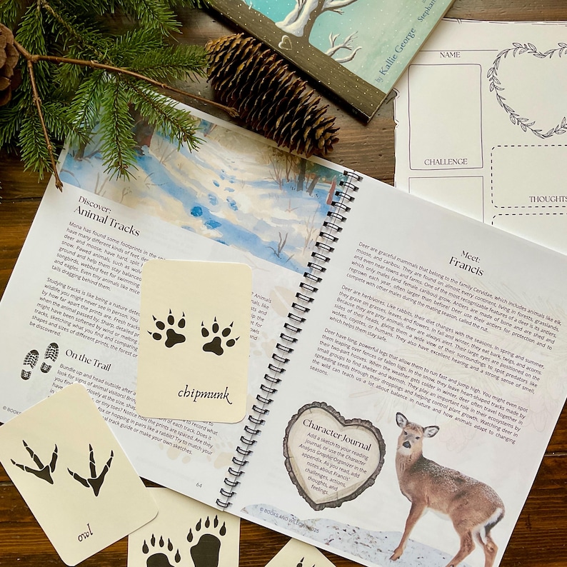 May include: An open nature journal with spiral binding, featuring illustrations of animal tracks and a deer. Includes cards with animal track illustrations and text, such as "chipmunk" and "owl". A pine cone and evergreen branch are in the background.