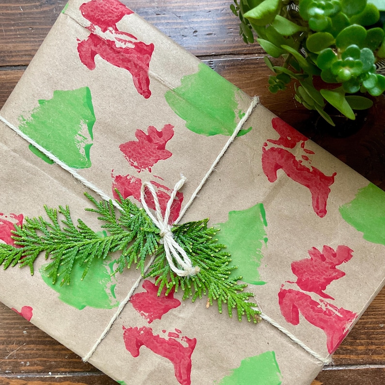 May include: A wrapped gift with brown paper featuring red and green painted designs. The package is tied with twine and adorned with a sprig of green foliage. The overall theme is festive and seasonal.