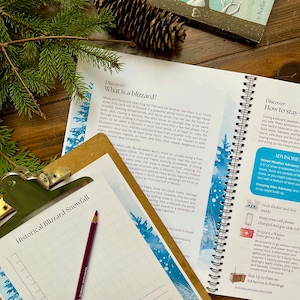 May include: An open book with the title "Discover: What is a blizzard?" and a clipboard with the title "Historical Blizzard Snowfall" are on a wooden surface. A pine cone and evergreen branches are in the background.
