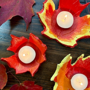 Puede incluir: Candelabros hechos a mano con forma de hoja en tonos vibrantes de rojo, naranja y amarillo, cada uno con una vela de t&eacute; blanca encendida. Los candelabros est&aacute;n dispuestos sobre una superficie de madera oscura, con hojas de oto&ntilde;o reales esparcidas.