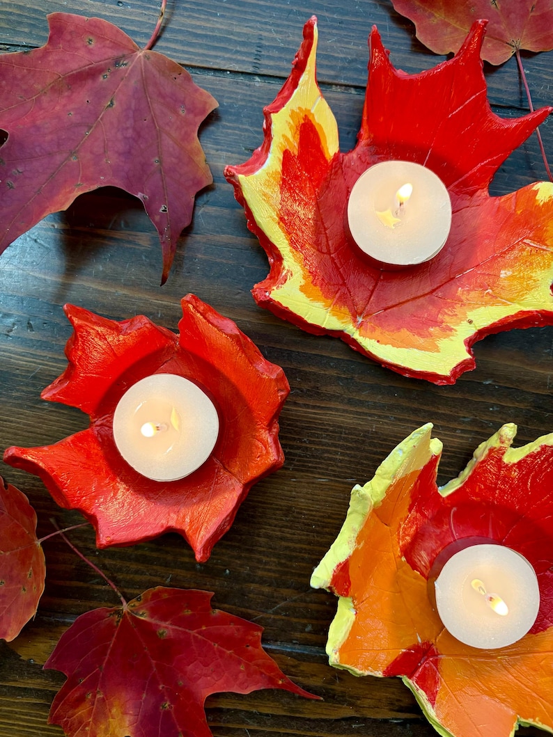 Puede incluir: Candelabros hechos a mano con forma de hoja en tonos vibrantes de rojo, naranja y amarillo, cada uno con una vela de t&eacute; blanca encendida. Los candelabros est&aacute;n dispuestos sobre una superficie de madera oscura, con hojas de oto&ntilde;o reales esparcidas.