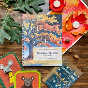 May include: A spiral-bound book titled "Heartwood Hotel" with an illustration of a tree with autumn foliage. The book is surrounded by fall-themed decorations, including leaf-shaped candle holders, acorns, and small art pieces featuring animal figures.