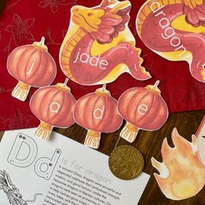 May include: A set of colorful felt cutouts for a Chinese New Year craft project. The cutouts include a red dragon, red lanterns with the letters 'j', 'a', 'd', 'e', and a gold coin. The text 'D is for dragon' is written on a white background.