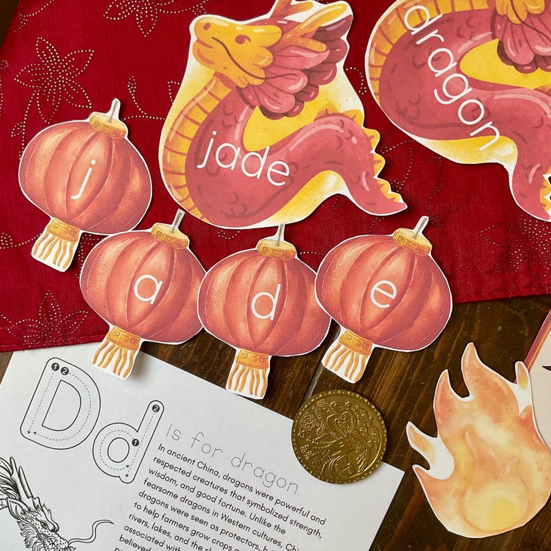 May include: A set of colorful felt cutouts for a Chinese New Year craft project. The cutouts include a red dragon, red lanterns with the letters 'j', 'a', 'd', 'e', and a gold coin. The text 'D is for dragon' is written on a white background.