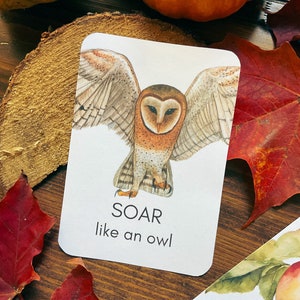 May include: A white card with a watercolor illustration of a barn owl with outstretched wings. The text "SOAR like an owl" is below the image. The card is on a wooden surface with fall leaves and pumpkins.