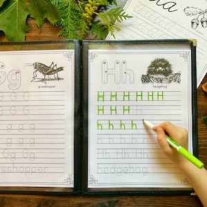 Pode incluir: Um caderno de exercícios em preto e branco com duas páginas mostrando as letras G e H. A letra G é ilustrada com um gafanhoto e a letra H é ilustrada com um ouriço. O caderno tem linhas para praticar a escrita das letras.