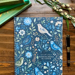 May include: A spiral-bound homeschool planner with a dark blue cover featuring a pattern of light blue and white birds, flowers, and foliage. The text "WILLOW GROVE HOMESCHOOL PLANNER" is printed in white on the front cover of the planner.