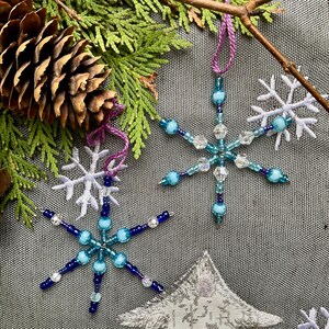 May include: Two beaded snowflake ornaments in shades of blue and clear, suspended by purple cords. The snowflakes are set against a backdrop of evergreen branches, a pine cone, and embroidered snowflake and tree shapes, suggesting a winter or holiday theme.