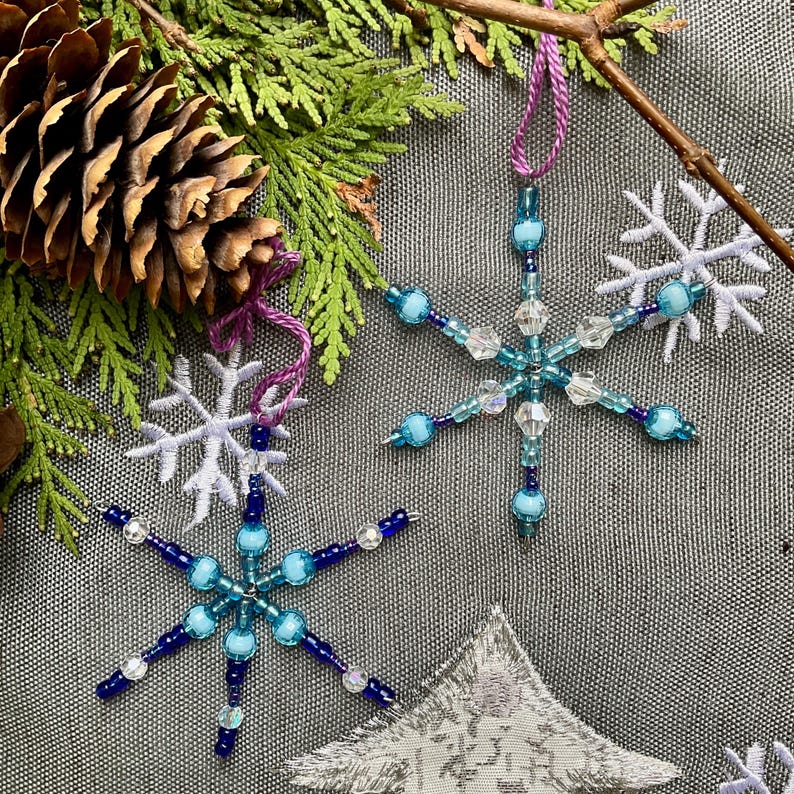 May include: Two beaded snowflake ornaments in shades of blue and clear, suspended by purple cords. The snowflakes are set against a backdrop of evergreen branches, a pine cone, and embroidered snowflake and tree shapes, suggesting a winter or holiday theme.