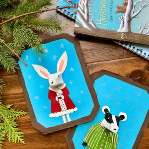May include: Handmade paper art featuring a bunny in a red coat and a cow in a green striped shirt, set against a blue background with snowflake accents. The artwork is framed with a brown border, and other notebooks are visible.