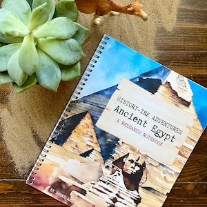 Puede incluir: Un cuaderno de notas azul y marrón con el título "History-Ink Adventures Ancient Egypt: A Research Notebook" y una imagen de un faraón egipcio y pirámides.