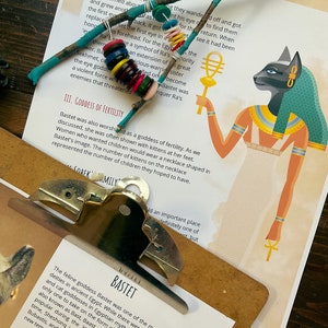 May include: Close-up of a page from a book about the Egyptian goddess Bastet. The page features a colorful illustration of Bastet, a cat-headed goddess, holding a staff. The text on the page discusses Bastet's role as a goddess of fertility and her association with cats.