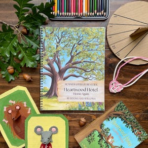 Könnte beinhalten: Ein spiralgebundener Sommer-Literaturführer mit dem Titel Heartwood Hotel Home Again von Books and Willows, der eine große Baumillustration zeigt, umgeben von Buntstiften, einer Sonnenuhr aus Pappe, einer rosa-weißen Kordel und zwei Maus-Bastelarbeiten.