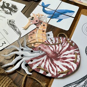 Op de afbeelding: Een set van vier papieren zeedieren met gaten om te rijgen. De zeedieren omvatten een zeepaardje, een nautilus, een octopus en een walvis. De zeedieren zijn bedrukt met aquarelachtige illustraties.