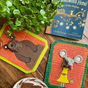 Puede incluir: Naturaleza muerta con un libro infantil titulado "Heartwood Hotel: A True Home" con una ilustraci&oacute;n de un rat&oacute;n. Dos obras de arte de cart&oacute;n hechas a mano representan un oso y un rat&oacute;n. Un plato con una porci&oacute;n de pastel de chocolate y follaje verde.