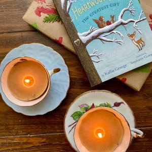 May include: Two lit candles in teacups with saucers, one light blue and the other white with floral designs. A children's book titled "Heartwood The Greatest Gift" and wrapped gifts are also in the image. The teacups and book are on a wooden surface.