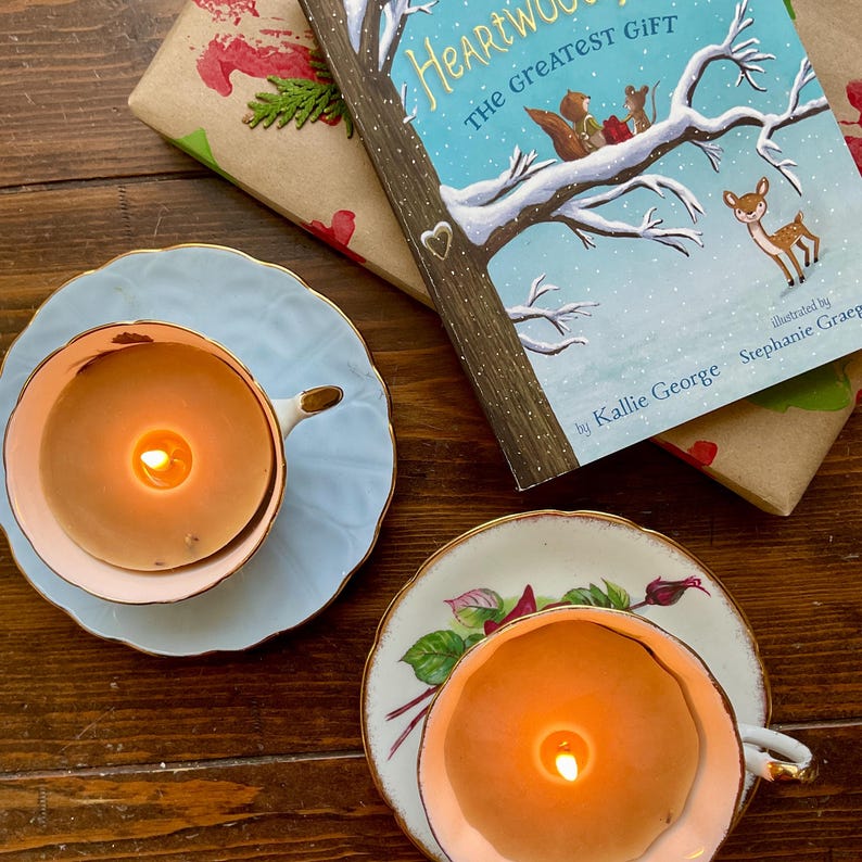 May include: Two lit candles in teacups with saucers, one light blue and the other white with floral designs. A children's book titled "Heartwood The Greatest Gift" and wrapped gifts are also in the image. The teacups and book are on a wooden surface.