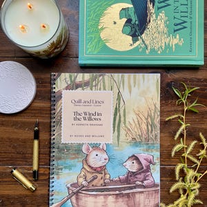 May include: A collection of items including a copy of "The Wind in the Willows" book, a spiral-bound notebook with the same title, a lit candle in a glass jar, a small white plate, and a gold pen. The book and notebook feature illustrations of the story.