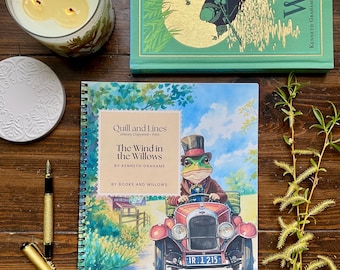 May include: A still-life arrangement featuring a lit candle in a glass jar, a decorative coaster, and a gold fountain pen. A spiral-bound book titled "The Wind in the Willows" is open, displaying an illustration of a frog driving a vintage car.