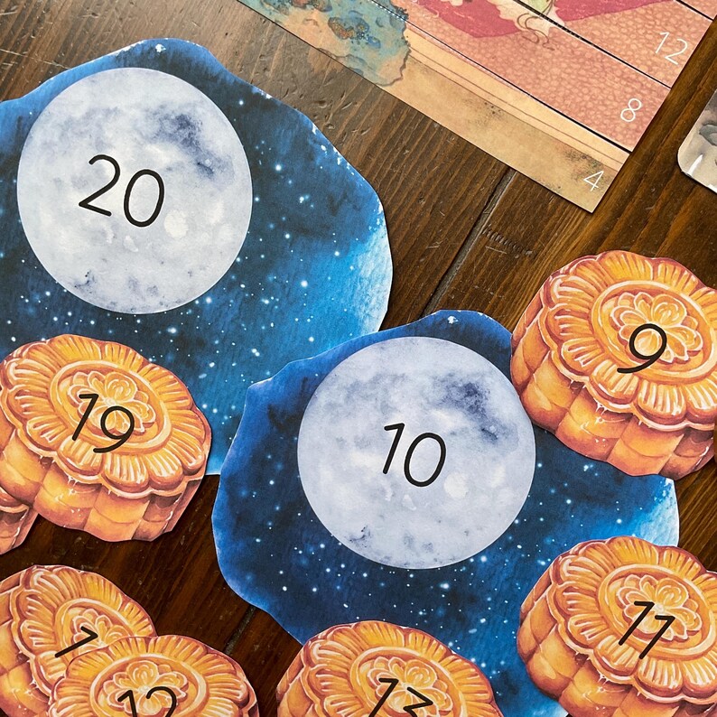 May include: A set of ten colorful moon and mooncake shaped paper cutouts with numbers 7, 9, 10, 12, 17, 19, 20, and 22 printed on them. The cutouts are against a wooden background.