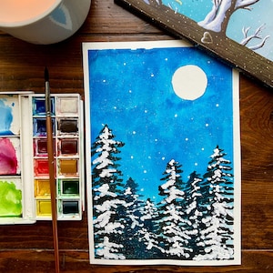 May include: Watercolor painting of a winter night scene with a full moon and snow-covered pine trees. A watercolor paint set and brush are next to the painting. A lit candle is in the upper left corner.