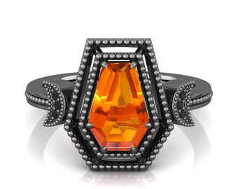 May include: A black ring featuring an orange gemstone in a coffin shape. The ring has a detailed frame around the gemstone and crescent moon accents on the sides. The ring is set against a white background.
